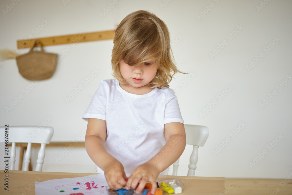 Creativity, early brain development and fine motor skills. Portrait of concentrated charming little 3 year old baby girl in white t-shirt playing with modeling clay or plasticine playdough at home Creativity, early brain development and fine motor skills. Portrait of concentrated charming little 3 year old baby girl in white t-shirt playing with modeling clay or plasticine playdough at home
