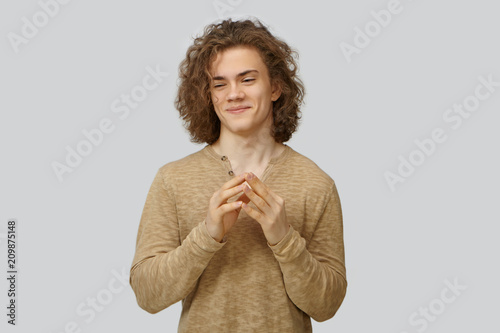 Studio shot of stylish sly young European man with voluminous curly hairstyle smiling mysteriously and clasping hands, scheming, planning tricky joke, going to prunk his best friend. Human emotions photo