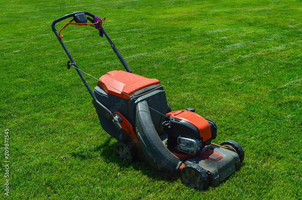 a large lawn mower is standing on the green grass. the device has already tonsured the lawn. gasoline lawn mower ready for use a large lawn mower is standing on the green grass. the device has already tonsured the lawn. gasoline lawn mower ready for use