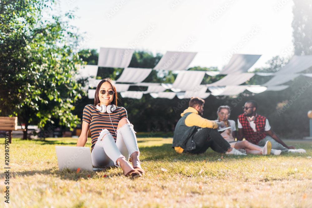 selective focus of young asian woman with laptop and multiracial friends behind resting on green grass in park selective focus of young asian woman with laptop and multiracial friends behind resting on green grass in park