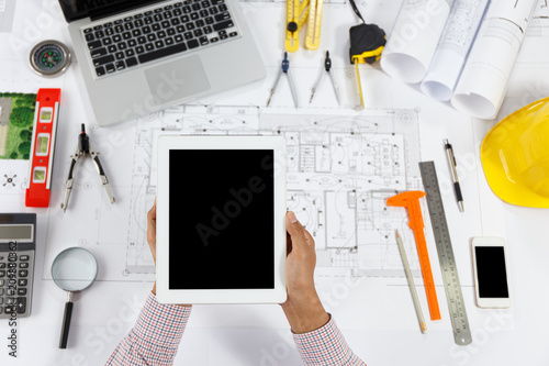 Top view, Architect or engineer hands using tablet working on blueprint plans with a pencil, ruler, calculator, smartphone, laptop and engineering tools. 