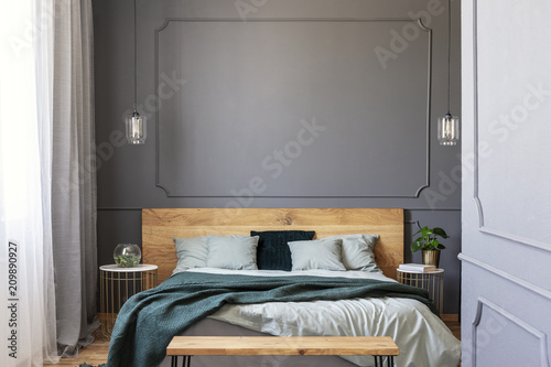 Green blanket on wooden bed with pillows in grey bedroom interior with wall with molding. Real photo
