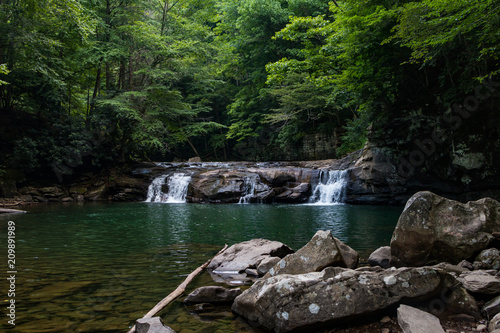 Large and wide waterfall flowing into a deep green pool, or swimming hole with large boulders.