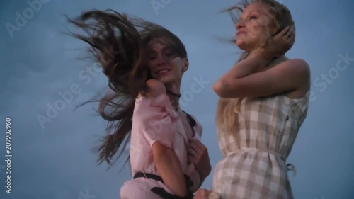Two young charming girls in dresses is enjoing wind, touching hair in field in rainy day in summer, freedom concept, flirt concept, bottom view
