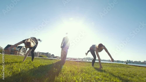 Young women performing acrobatic wheel on the grass, silhouette of young man standing in center of them