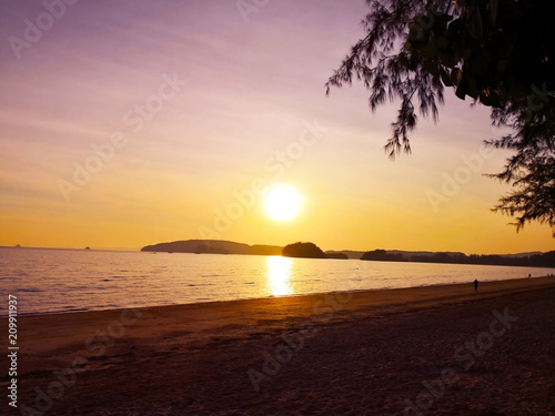 In the summer vacation. Silhouette of people are walking on the tropical bay beach, where is full of pine trees. As the sun is falling down behind the island. Selective focus and copy space.