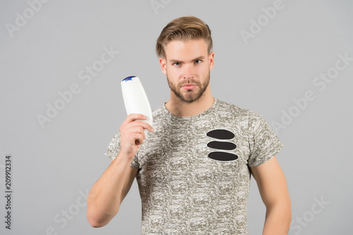 Man strict face holds shampoo bottle, grey background. Man enjoy freshness after washing hair with shampoo. Hair care and beauty supplies concept. Guy with hairstyle holds bottle shampoo, copy space photo
