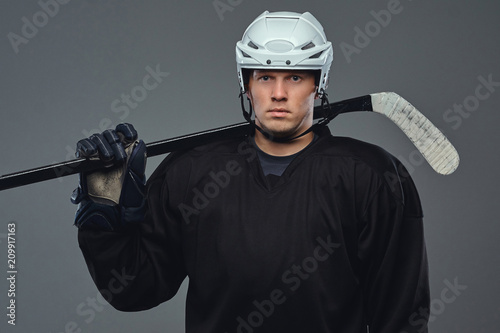 Hockey player wearing black protective gear and white helmet holds a hockey stick on a gray background.