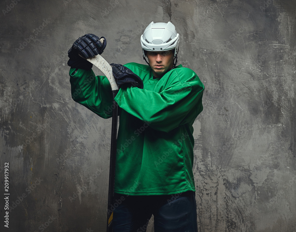 Hockey player wearing green protective gear and white helmet standing with the hockey stick on a gray background. Hockey player wearing green protective gear and white helmet standing with the hockey stick on a gray background.