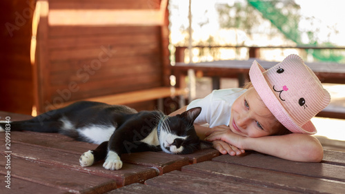 Cute little girl in pink hat petting a black cat outdoors.