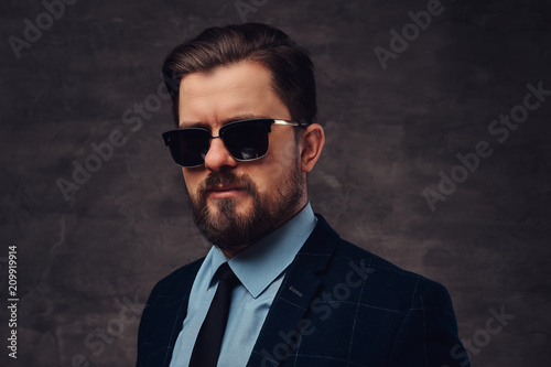 Close-up portrait of a handsome fashionable middle-aged man with beard and hairstyle dressed in an elegant formal suit and sunglasses. Isolated on a textured dark background in studio.