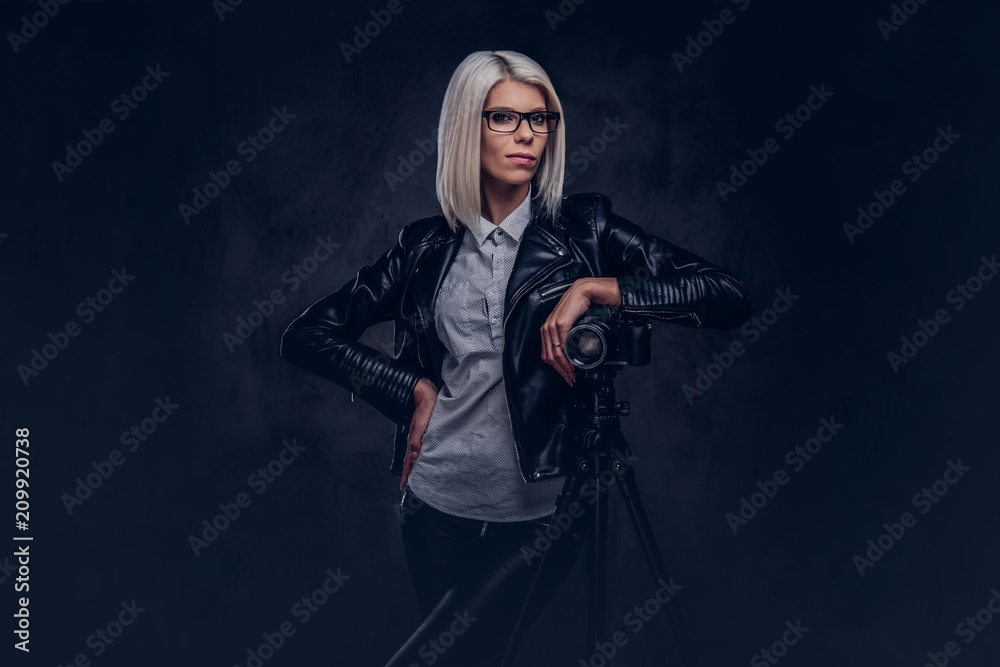 Smart blonde female photographer in trendy clothes posing while leaning on a professional camera with a tripod at a studio, looks at the camera.