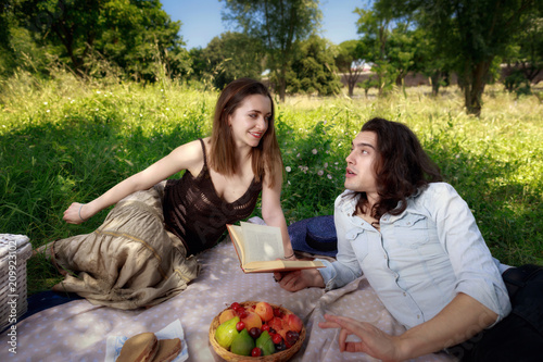 A couple of young people, man and woman, lying in the park grass at a picnic. He reads some verses from a book while she listens to him.
