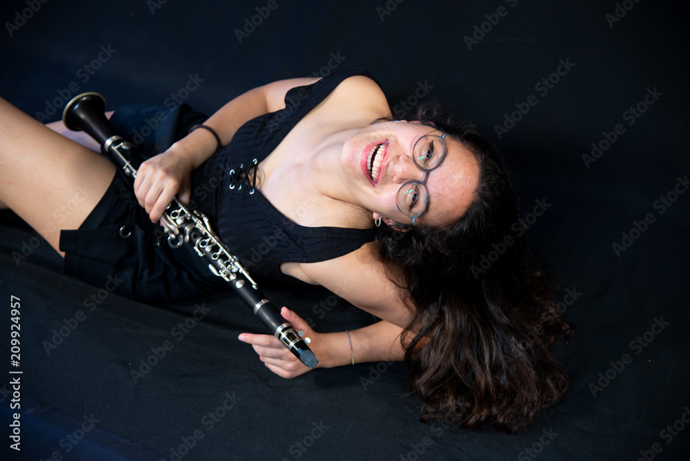 A brunette girl, with glasses, lying laughing and holding her clarinet music instrument isolated on a black background