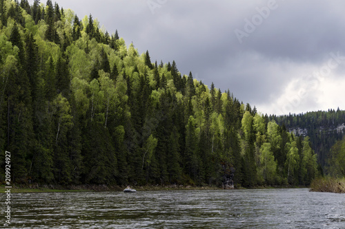 landscape of the Ural river Usva, the Chusovaya tributary, with picturesque wooded coastal cliffs and a fishing inflatable boat in the distance, on a cloudy day,