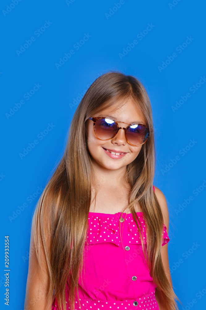 Portrait of young pretty girl in pink dress, sunglasses and hat on blue background . Summer vacation and travel concept Portrait of young pretty girl in pink dress, sunglasses and hat on blue background . Summer vacation and travel concept
