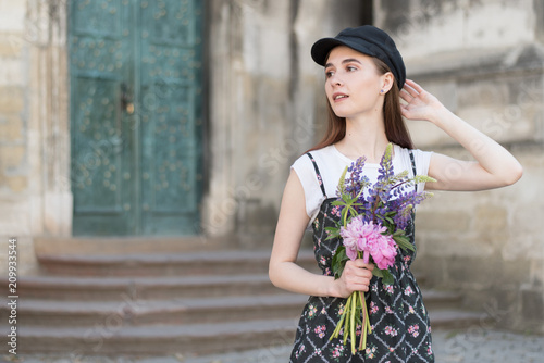 Playful girl with perfect long hairs posing outdoor. Wearing wool cap. Street fashion look. Cute teenage girl with flowers. Beauty and fashion summer concept. Model. Walk in the city