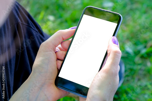 The girl is sitting on the grass and looking into the smartphone holding it in her hands. Mockup image.