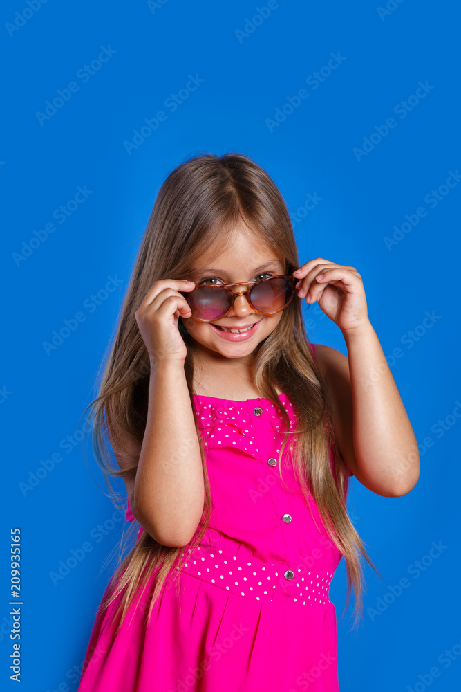 Portrait of young pretty girl in pink dress, sunglasses and hat on blue background . Summer vacation and travel concept Portrait of young pretty girl in pink dress, sunglasses and hat on blue background . Summer vacation and travel concept