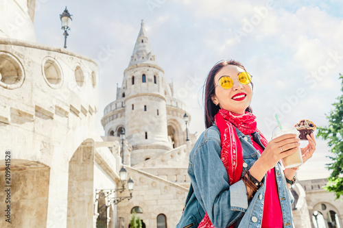 Young asian woman tourist drinking frappe sweet cold coffee and tasty muffin at the Budapest city street near Fisherman Bastion. Travel in Hungary and fast food break concept