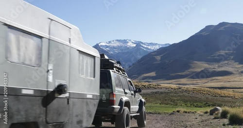 Van with trailer driving on gravel road, passes through badoo, stream. Surrounding of Varvarco valley, mountains with snow at background. Camera stays still while motorhome moves. Patagonia photo