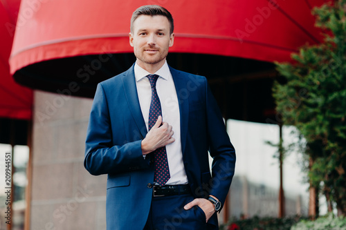Handsome male company owner dressed formally, stands outside, has confident expression, ready for business meeting. Successful young attractive executive manager poses outdoor near office building