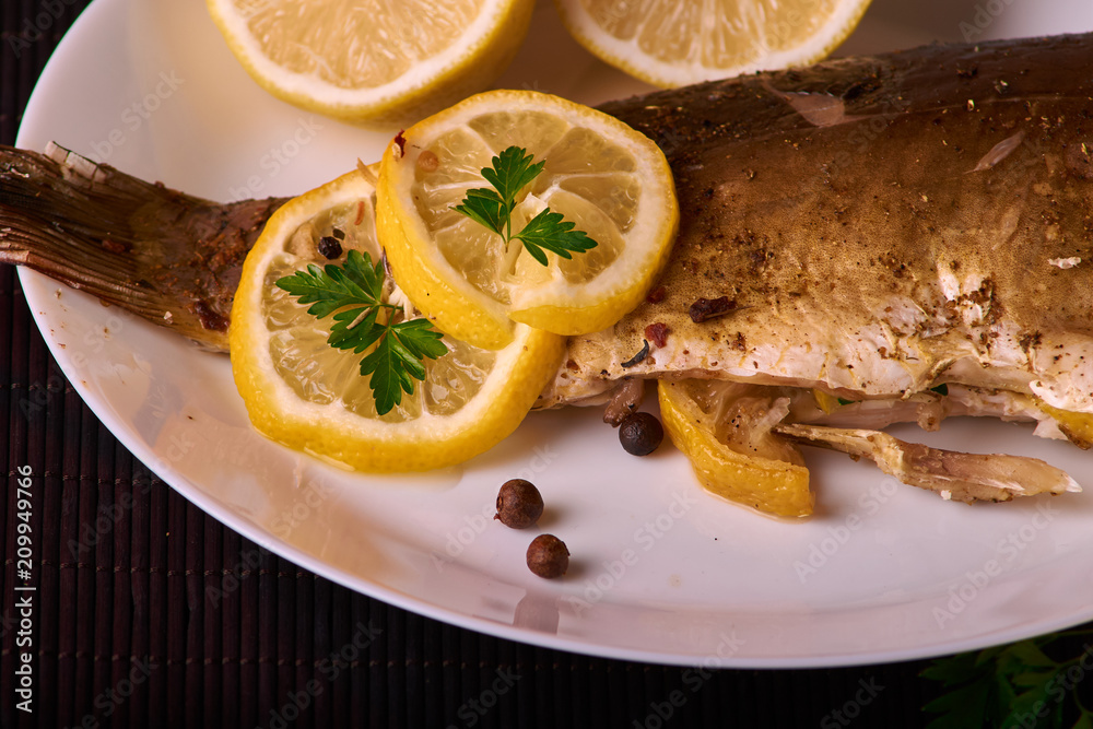 Baked fish with lemon, spices and herbs on white plate on wooden background without head Baked fish with lemon, spices and herbs on white plate on wooden background without head