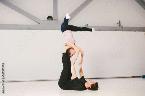 A young Caucasian male and female couple practicing acrobatic yoga in a white gym on mats. They are in the STAR pose
