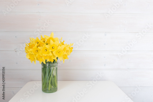Fototapeta Naklejka Na Ścianę i Meble -  Yellow daffodils in glass jar on white table against rustic white wood panelled wall with copy space