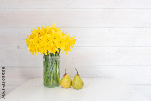 Fototapeta Naklejka Na Ścianę i Meble -  Yellow daffodils in glass jar on white table with two green pears against rustic white wood panelled wall