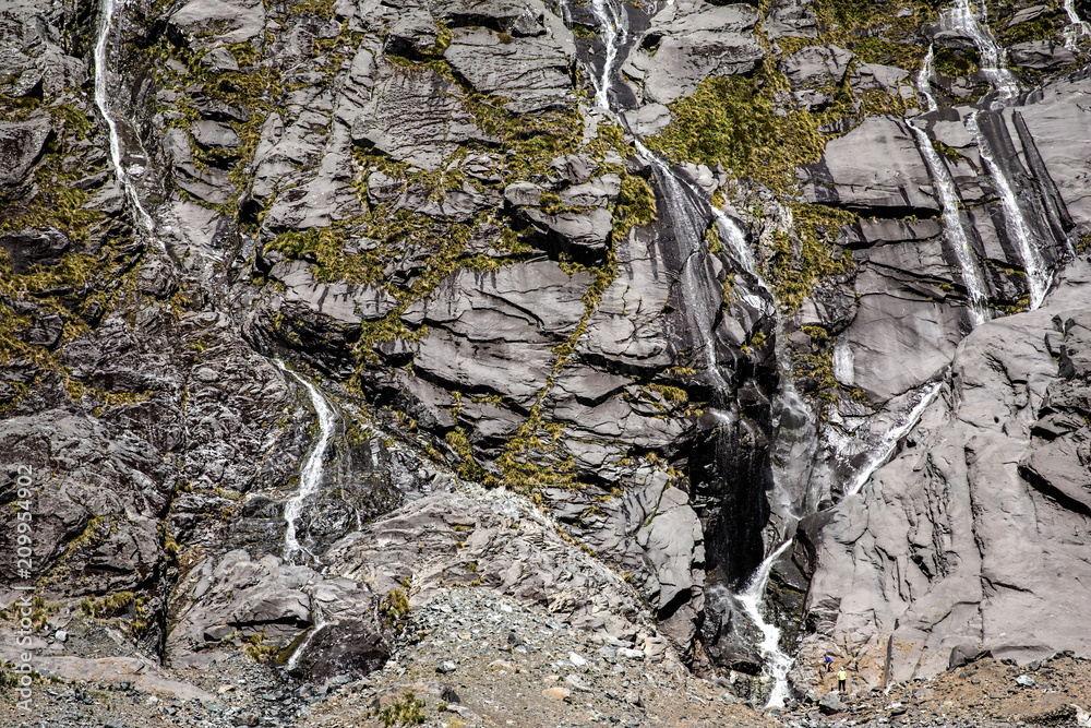 Hikers admiring multiple waterfalls on a cliff face on the road to Milford Sound, South Island, New Zealand