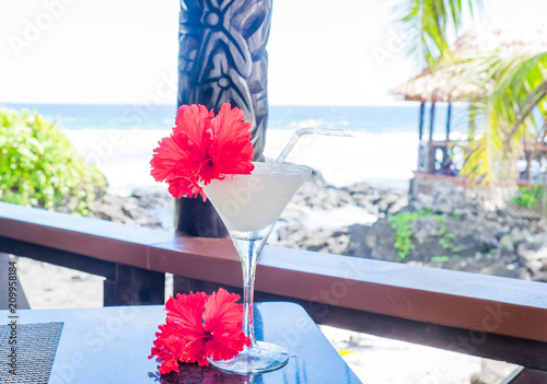 Alcoholic cocktail drink with red hibiscus flower garnish with wooden carving in background at beach bar on Upolu Island, Samoa, South Pacific photo