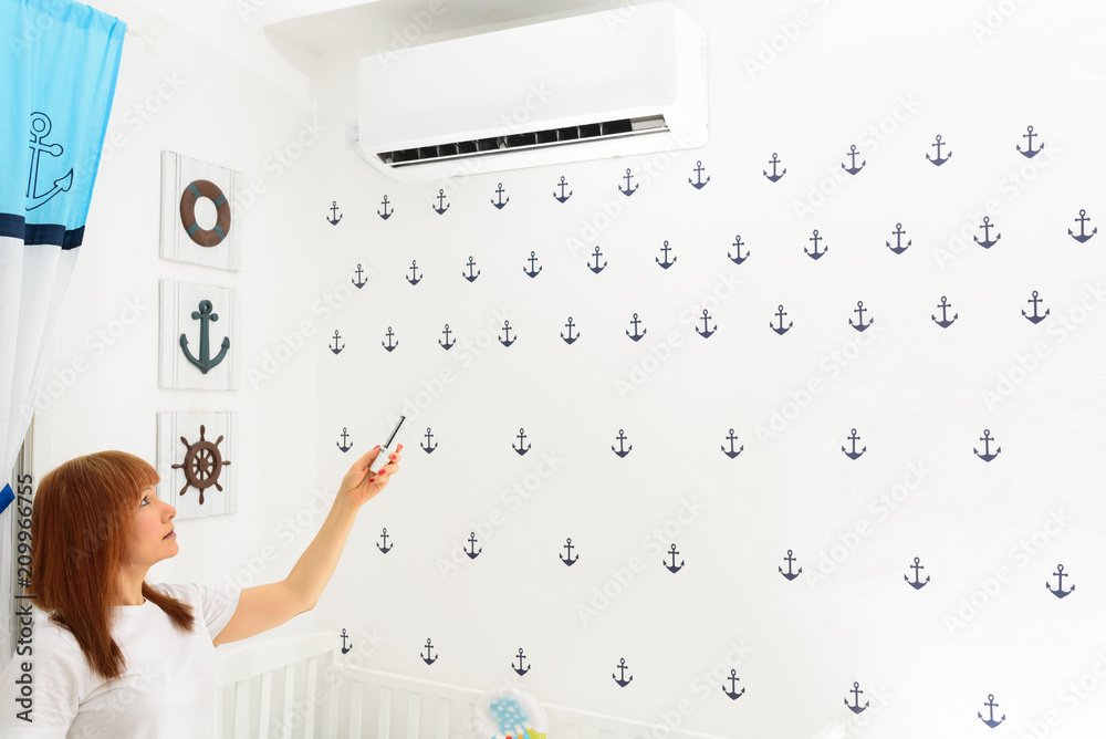 Obraz premium Portrait Of A Happy Woman Holding Remote Control In Front Of Air Conditioner At Home in childrens room.Air conditioner inside the cute baby boy’s room.With hot weather,is a good solution for family.