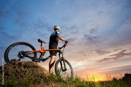 Cyclist standing alone with orange bicycle and holding hands on handle bar. Athletic man wearing black sportswear and helmet and posing on hill. Concept of healthy lifestyle and sport activities.
