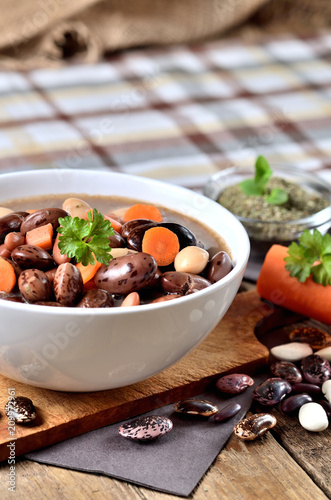Bowl of bean soup with large beans on cutting board, carrots, parsley, marjoram, spoon and ladle, towel in background - vertical photo