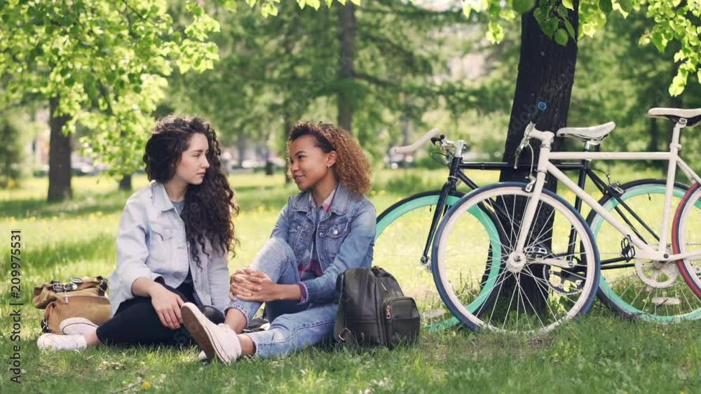 Cheerful young women Caucasian and African American are chatting in park while resting after riding bikes in beautiful green park. Girls are careless and happy. Cheerful young women Caucasian and African American are chatting in park while resting after riding bikes in beautiful green park. Girls are careless and happy.