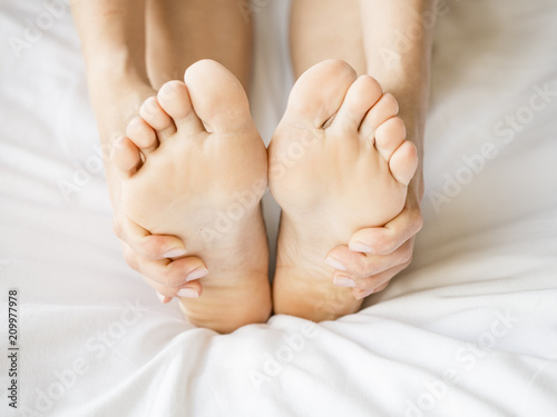 Woman Holding her Tired Feet in Hand Sitting on Bed with White Sheets. © Stavros