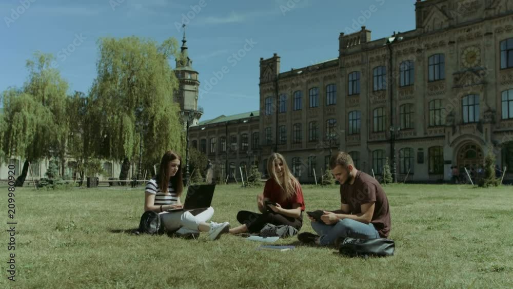 Cheerful college students studying with electronic devices and discussing university project while sitting cross-legged on campus lawn. High school classmates networking with digital devices on lawn. Cheerful college students studying with electronic devices and discussing university project while sitting cross-legged on campus lawn. High school classmates networking with digital devices on lawn.