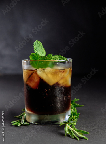 Glass of cola with ice, mint and lemon, rosemary on black background