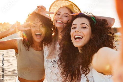Three joyful multiethnic women 20s in summer clothing smiling at camera, while taking selfie photo during holiday on nature photo
