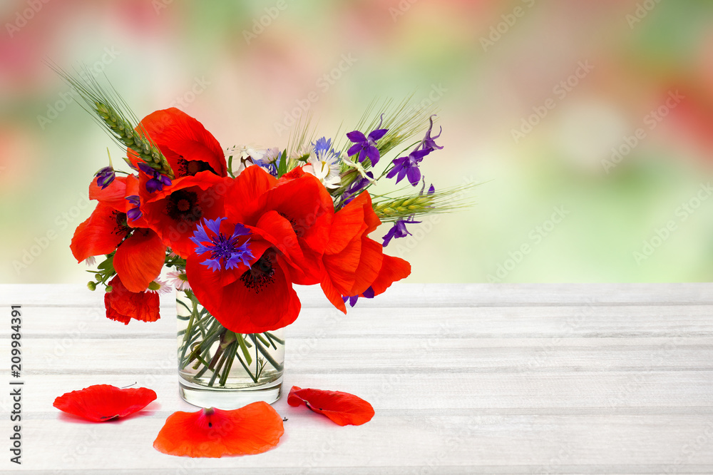 Obraz premium Red poppies, cornflowers, ears of green wheat, chamomile in small vase on white wooden table on summer blur natural background with space for text.