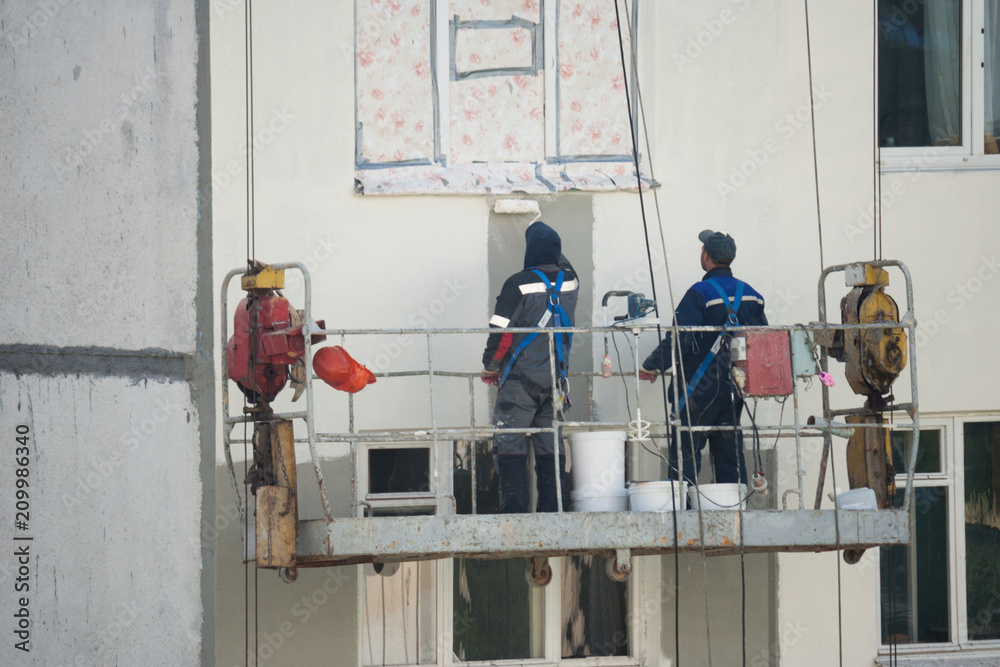 Two plasterers, the painter perform painting on the facade of the building at the height of the building cradle. A view of the job of painting the wall from the back of the workers.
