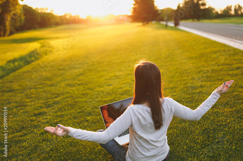 Young calm student female in casual clothes. Woman sitting, relaxing, hands in yoga gesture. Laptop pc computer on green grass ground sunshine lawn outdoors. Mobile Office. Freelance business concept.