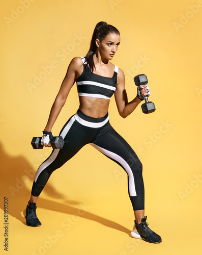 Attractive strong woman working with dumbbells. Photo of sporty latin woman in fashionable sportswear on yellow background. Strength and motivation.