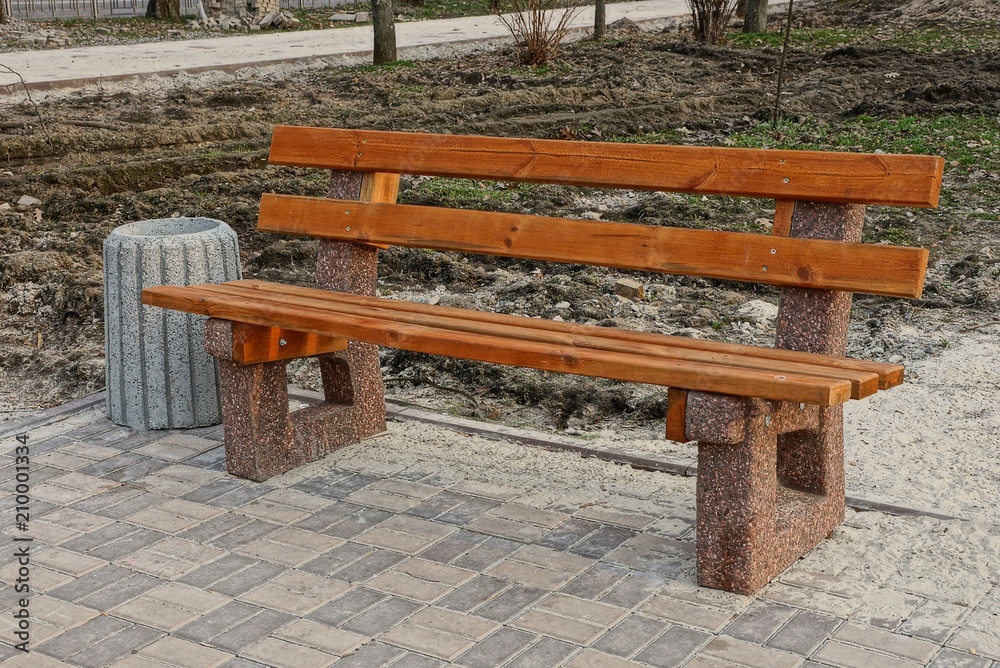 brown wooden bench and a gray urn on the sidewalk in the park brown wooden bench and a gray urn on the sidewalk in the park