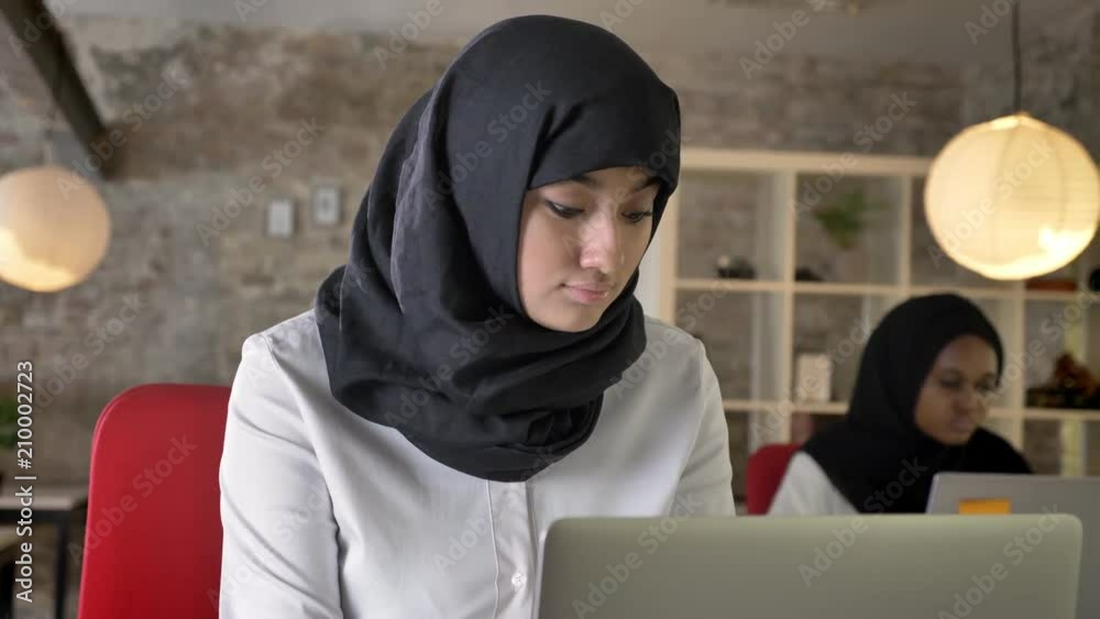 Young muslim women in hijab working and looking into camera, smiling, two womens sitting in office, one of them black women in hijab Young muslim women in hijab working and looking into camera, smiling, two womens sitting in office, one of them black women in hijab