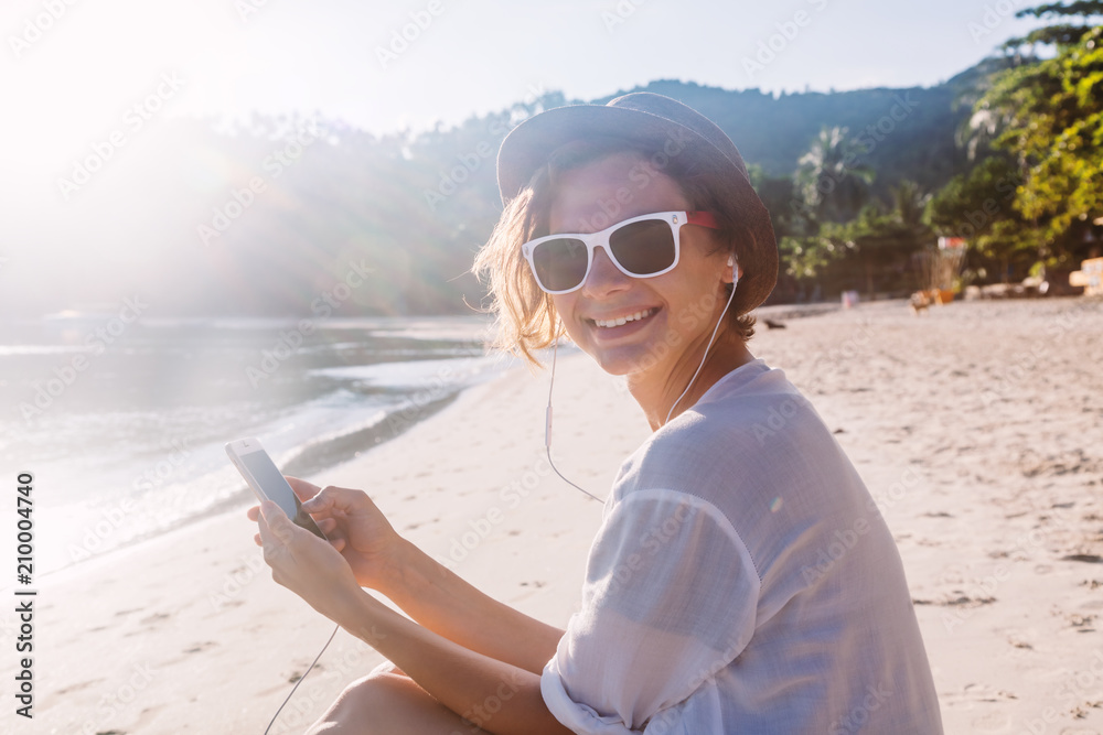 Young beautiful woman hipster traveler in headphones and with mobile phone in hands, on the beach in the sun
