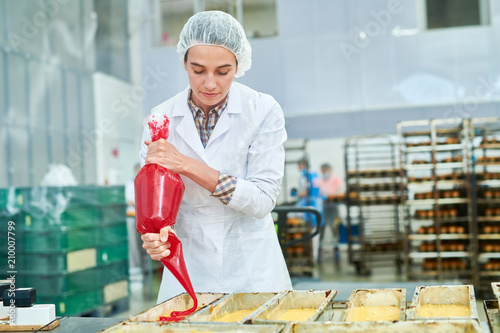 Confectionery factory employee in white coat pouring red cream from pastry bag.  photo