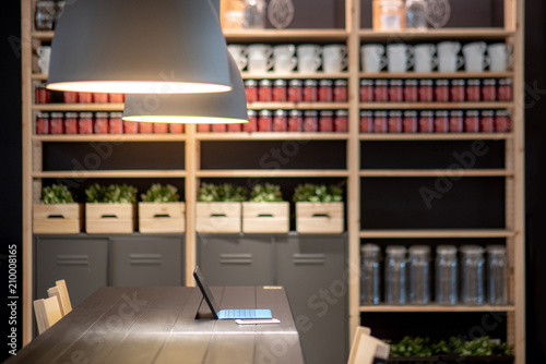 Digital tablet and smartphone on wooden table desk in working area with modern lamps overhead and decoration shelves in the background. Coworking space or design office workspace concept.