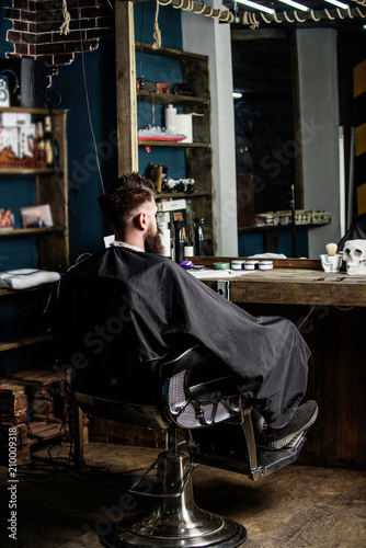 Man with beard client of hipster barbershop. Hipster with beard waits for barber and haircut. Man with beard covered with black cape sits in hairdressers chair in front of mirror. Barbershop concept